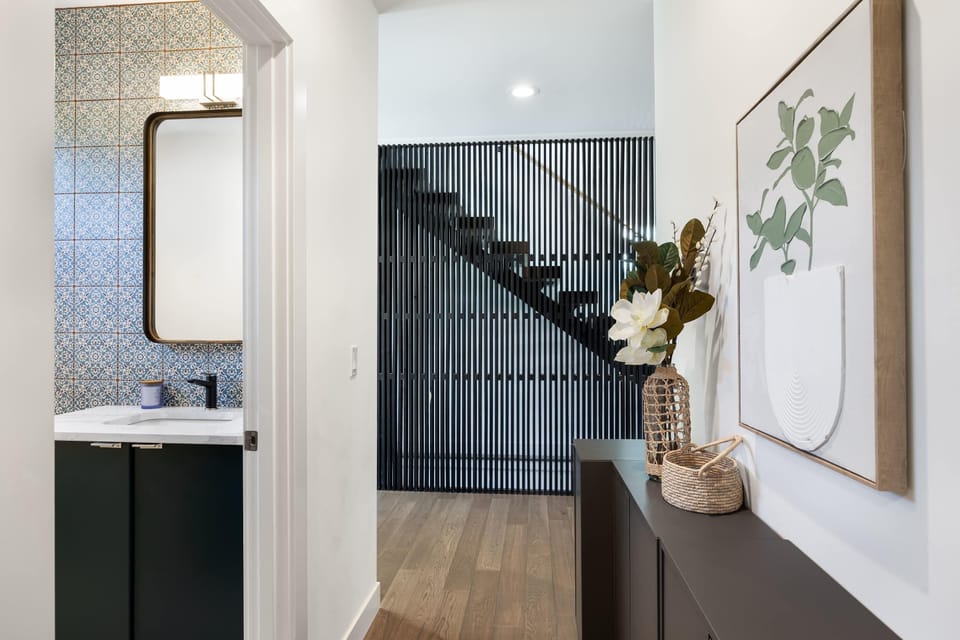 Console table displays fresh flowers beneath plant artwork,leading to bathroom and stairway slats.