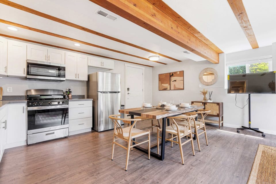 Modern open-concept kitchen-dining area​ featuring white cabinetry with integrated stainless appliances, a central wood-grain dining table, and thoughtful details like a mobile TV stand.