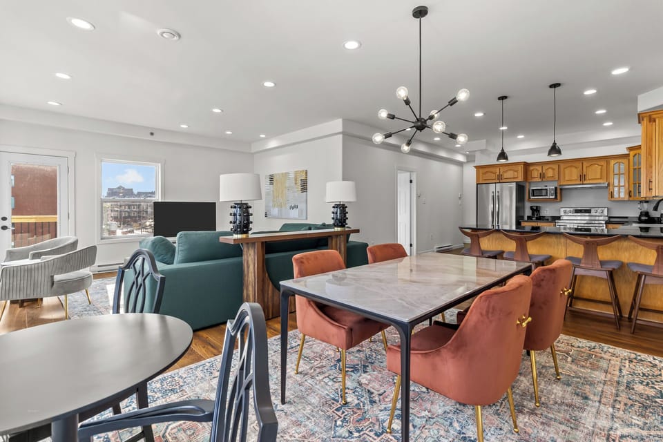 "Modern open-concept living area with a stylish dining table, plush orange chairs, cozy green sofa, and sleek kitchen. Well-lit with recessed lighting and a contemporary chandelier. Perfect for relaxation and entertaining.