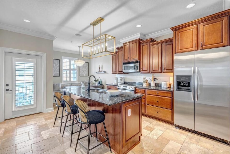 - Stylish kitchen with granite island, wood cabinets, and stainless fridge
- Bar seating for three under a gold light fixture adds flair and function
- Natural light pours in from patio door and shuttered windows nearby