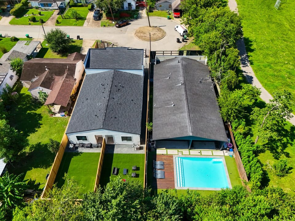 An aerial view of two modern adjacent properties, one with a large fenced backyard and the other featuring a swimming pool with lounge chairs.