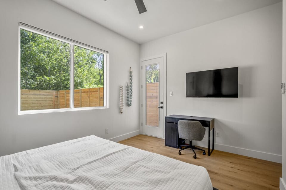 A bright bedroom with a large window, wall-mounted TV, small desk with a chair, and a door leading to an outdoor space.