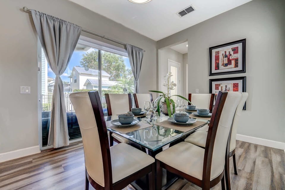 Dining area with glass table and cushioned chairs next to a large window with gray curtains