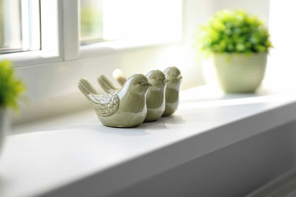 A windowsill with a ceramic bird decoration. A potted plant in the background adds freshness to the room.