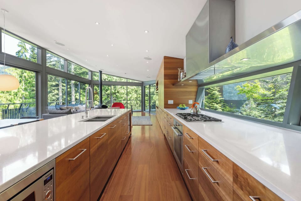 - Long kitchen layout with dual counters and full lighting  
- Expansive windows framing vibrant greenery outside  
- A chef’s dream for cooking and conversation combined  
