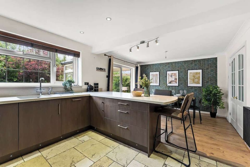 The modern kitchen features a breakfast bar with stylish high-back stools, overlooking the dining area and garden beyond.