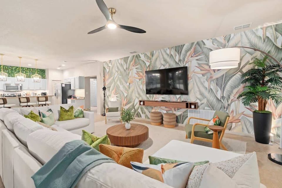Spacious Orlando family villa living room with tropical leaf feature wall, large white sectional sofa, smart TV, rattan accent chair, ceiling fan, and lush potted plants—perfect vacation rental lounge near Disney and Universal.