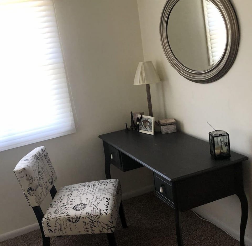 This serene master bedroom features a cozy workspace with a dark wood desk, patterned chair, and soft neutral tones. A round mirror and window with natural light enhance the calm ambiance, creating a perfect retreat for rest and relaxation.