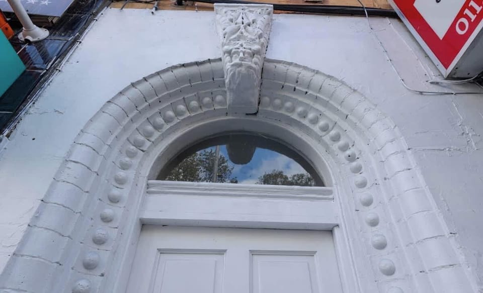 A white, ornate arched doorway with intricate detailing above a double door numbered 48A. The door features a semi-circular window and is part of a classic architectural facade. The photo is taken from a low angle.