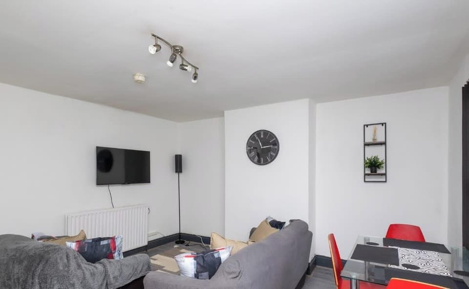 A cozy, modern living room with a grey sofa, wall-mounted TV, and a dining table with red chairs. The space is bright and inviting, ideal for relaxing or entertaining in a comfortable home setting.