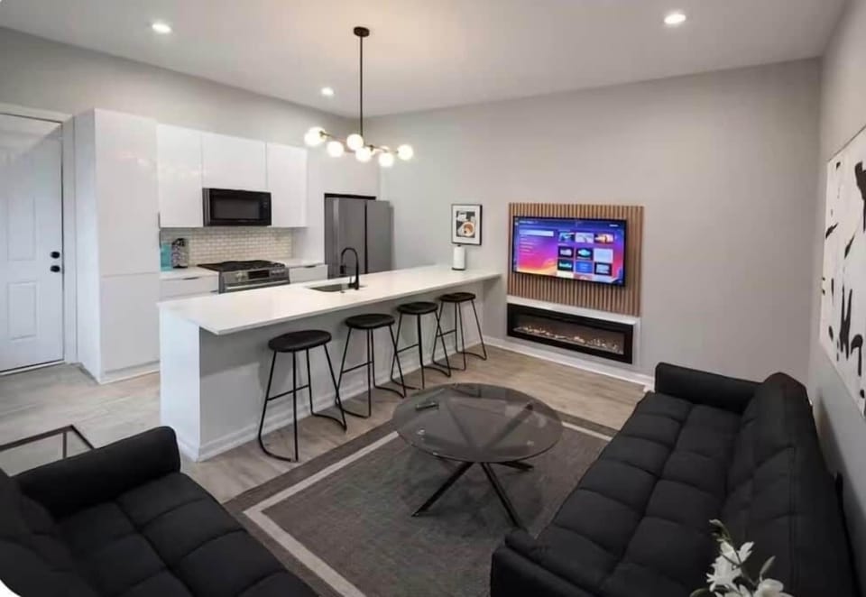 Stylish  living space featuring two futon sofa beds, a flat-screen TV, and a modern electric fireplace. The large island bar offers generous seating and flows into the kitchen, creating a comfortable space to relax, dine, or entertain.
