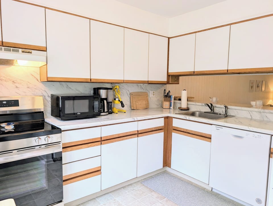 718D Treetop Lane - Kitchen Workspace - The kitchen features a practical U-shaped layout with white cabinetry accented by natural wood trim. The space is well-equipped with modern appliances, including a stainless steel gas range, a black microwave, and a white dishwasher, all set against neutral countertops.