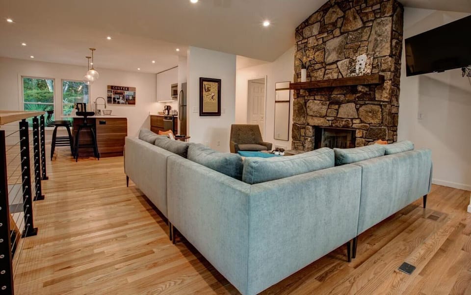 🔥 Inviting living space with a large sectional sofa 🛋️, rustic stone fireplace 🌿, and open layout leading to a modern kitchen ☕—perfect for cozy nights and social gatherings 🍷.