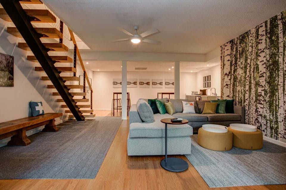 🛋️ Spacious lounge area with a modern sectional sofa, colorful pillows 🌈, and a striking birch tree mural 🌿, plus open stairs and stylish lighting—perfect for relaxing or entertaining 🍷.