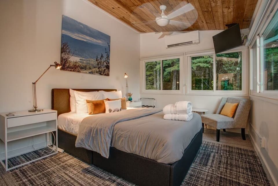 🛏️ Inviting bedroom with a king bed, warm accent pillows 🧡, cozy seating nook 🪑, and large windows framing peaceful forest views 🌲—perfect for restful nights.
