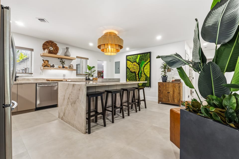 A wide, glowing view from kitchen to living room—where the waterfall-edge quartzite island that seats 6, lush plants, and smooth cement floors meet a handwoven Tulum chandelier and signature Tabu Collection pieces. Warm, connected, and curated.