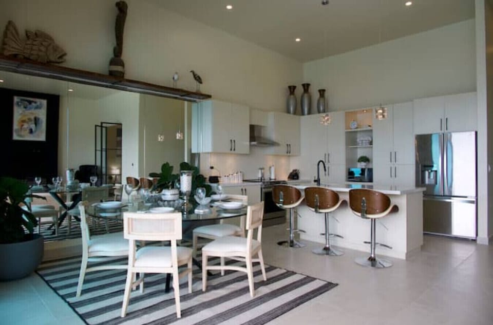 Bright, open kitchen and dining space with high ceilings, sleek white cabinetry, bar seating, and a glass-top table. Clean lines and natural light complete the look.