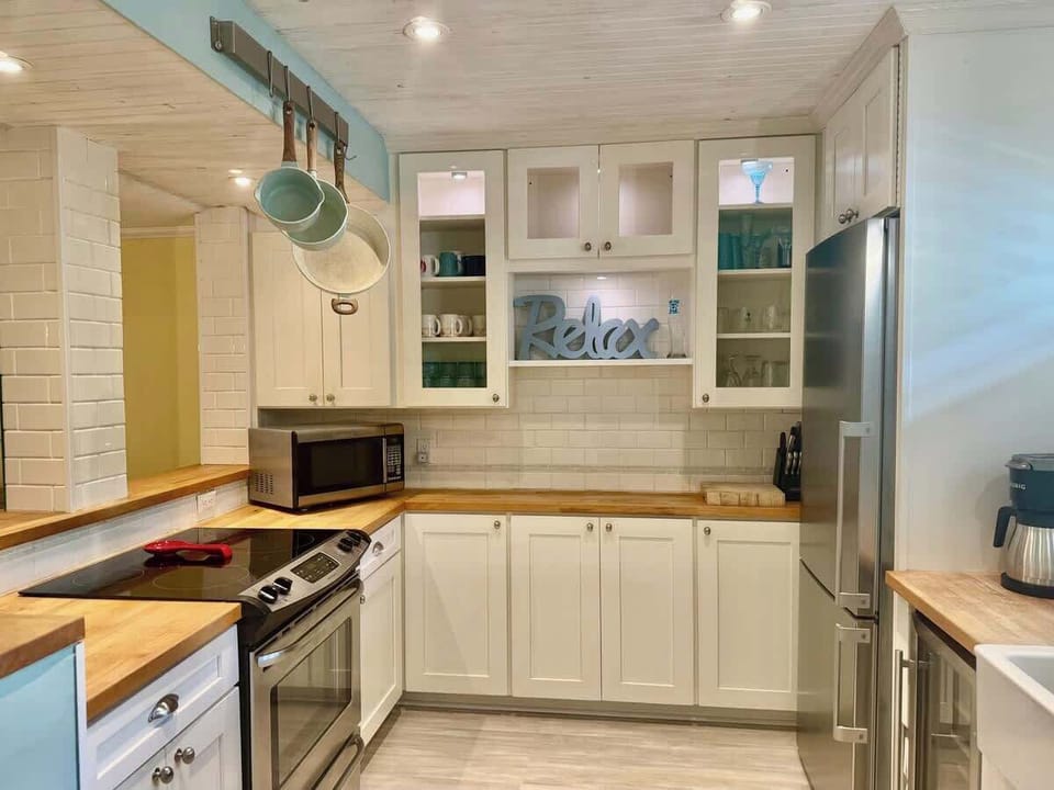 A bright, clean kitchen featuring white cabinetry, warm wood countertops, and high-end stainless steel appliances. The space is well-equipped with a deep farmhouse sink, a built-in wine cooler, and plenty of storage.

