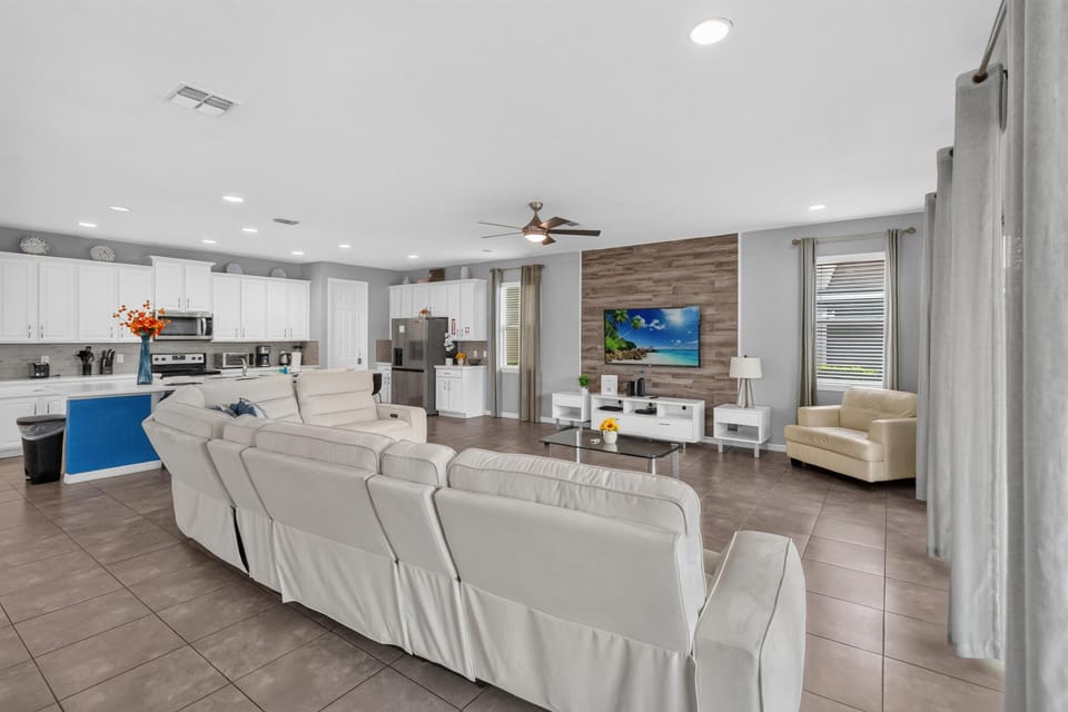Open-concept living area with white leather sectional, wood-paneled TV wall, and gray tile floors throughout.