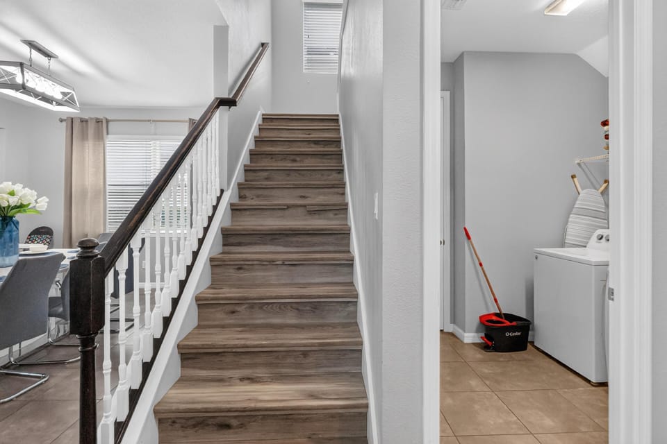 Wood-look stairs with dark handrail lead upstairs beside a laundry room with white washer, iron board, and tan tile floor.