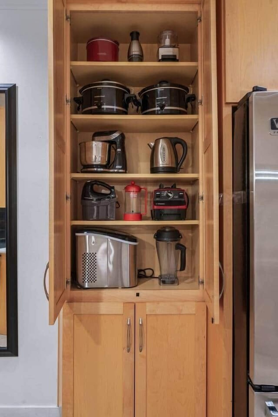 A well-organized kitchen cabinet showcasing a variety of appliances, including slow cookers, kettles, blenders, and a toaster, neatly arranged beside a stainless steel refrigerator.