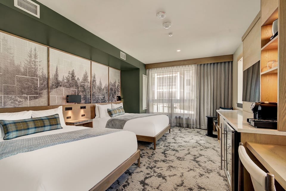 Sheer-curtained window, two queens, and a sitting chair — accessible spacing that still reads as a proper mountain room