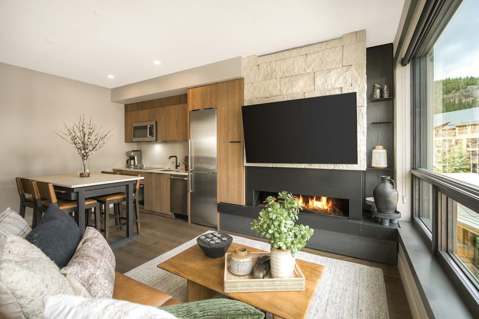 Open kitchen and living space with stone fireplace and wide windows letting in natural light