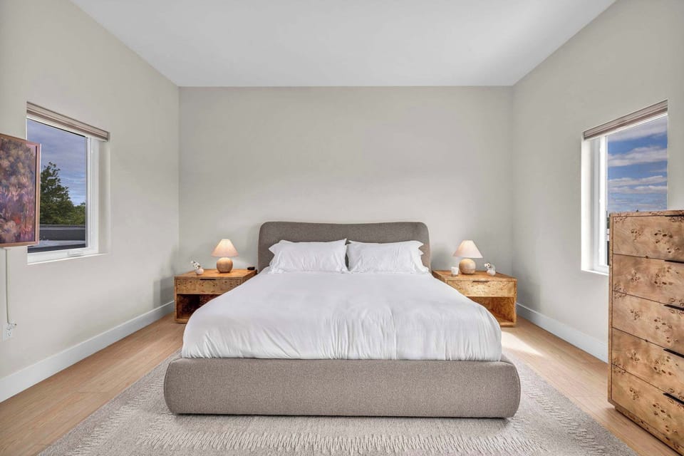 Wake up refreshed in a calming bedroom with a plush king bed, cozy lighting, and natural light from two windows for the perfect start to your day.
