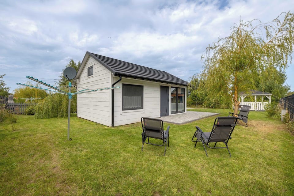 A small white cottage with dark roofing, surrounded by a green lawn. Outdoor chairs and a table create a relaxing garden space.