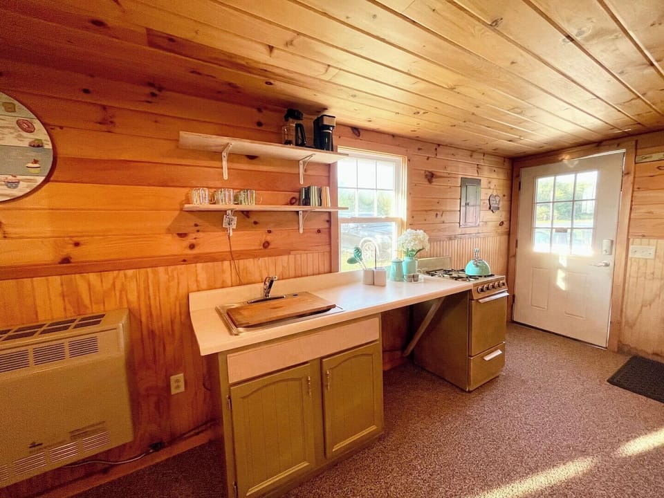 Functional kitchen with open shelves, countertop workspace, and a compact stove for preparing meals. Rustic wood-paneled walls add to the cabin charm. Mini fridge available for use.