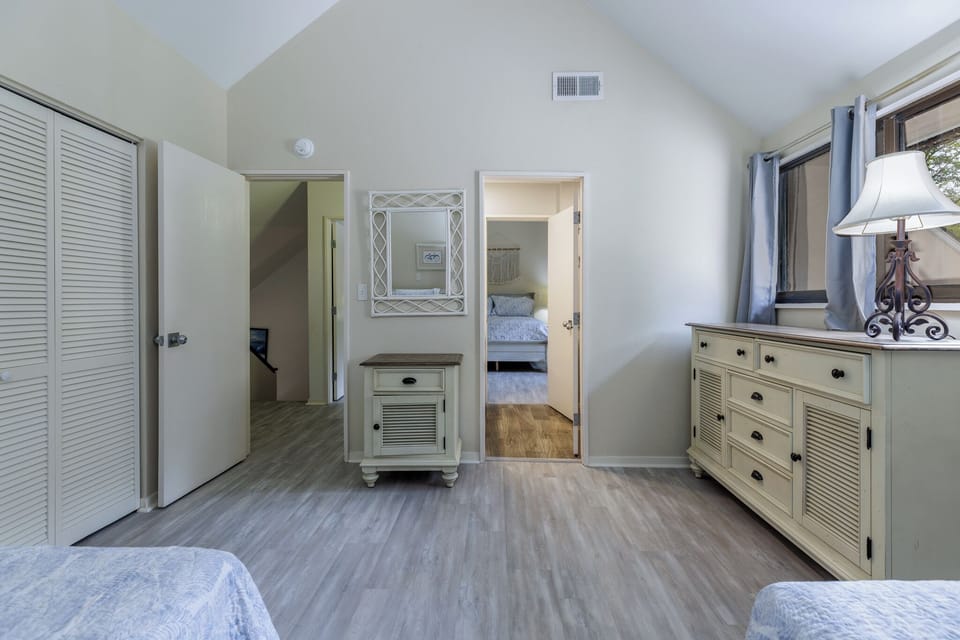 Cozy bedroom with light gray floors, cream furniture, and soft blue accents, featuring a dresser, mirror, and views into an adjoining room with a neatly made bed.