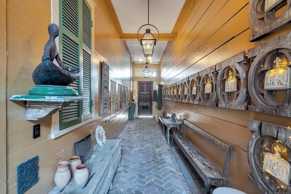 From the garden door, the passageway stretches toward the front entrance, lined with rustic benches and illuminated religious plaques set into carved wooden frames. Warm gold walls, lantern lighting, and brick floors enhance the historic charm.