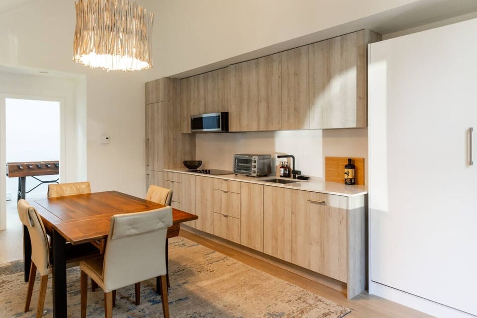 This modern kitchen features light wood cabinetry, a built-in fridge, stovetop, microwave, toaster oven, and essentials. A wood dining table with padded chairs sits under a stylish light fixture, with a foosball table nearby for added fun.