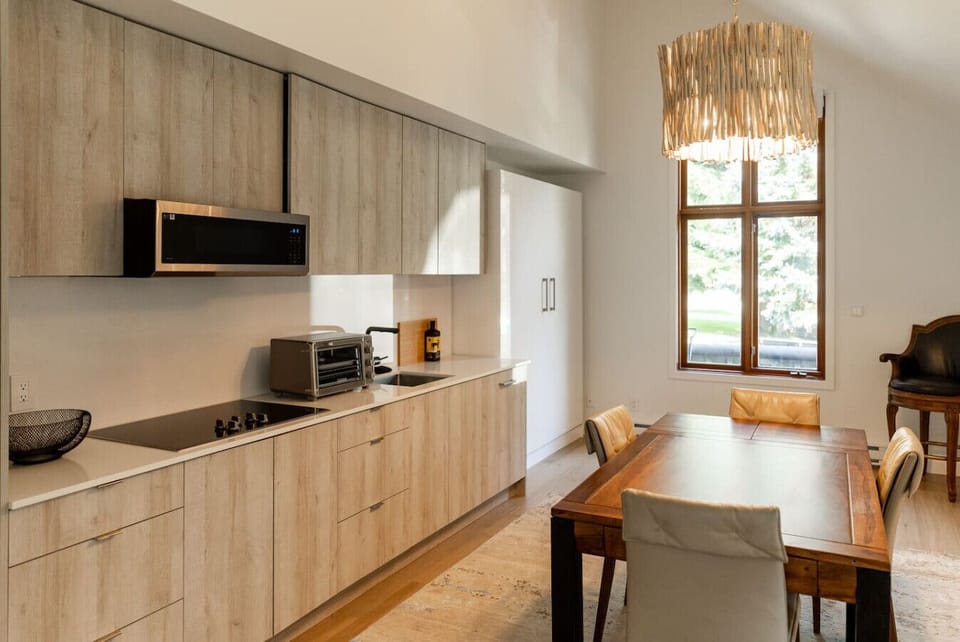 This modern kitchen features light wood cabinetry, a built-in fridge, stovetop, microwave, toaster oven, and all the essentials. A wood dining table with padded chairs sits beneath a stylish light fixture, creating a warm and inviting dining space.