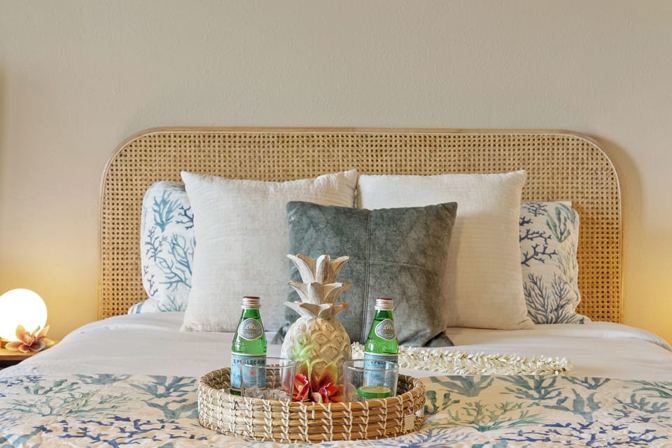 Close‑up of the bed with wicker headboard, decorative pillows and basket filled with bottled drinks, pineapple and tropical flowers adding a welcoming touch.