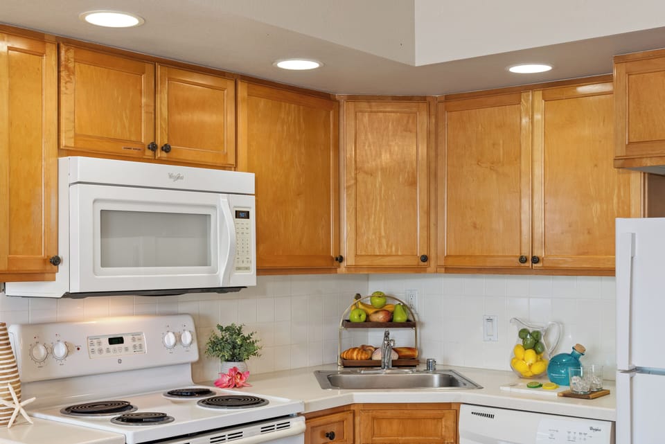 Another kitchen view with similar cabinetry and appliances, recessed ceiling lights brightening the space, glass‑top table with fruit bowl nearby.