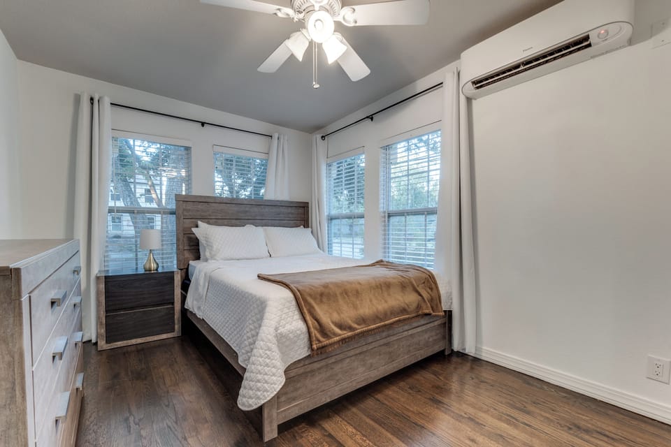 Rest peacefully in your comfortable bedroom with crisp white linens, warm wood furnishings, and abundant natural light from large windows.