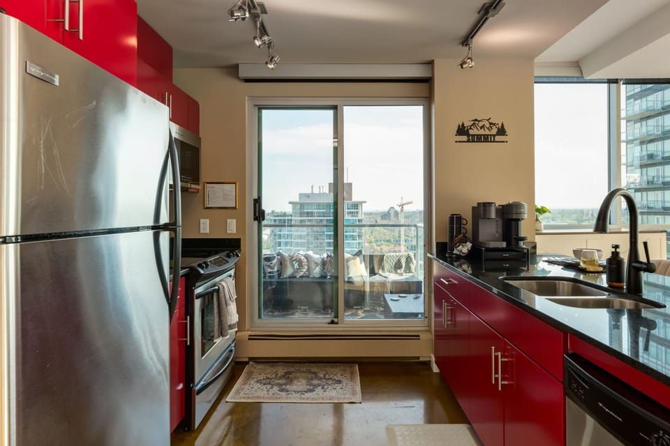 Stylish kitchen with bold red cabinets, black countertops, and stainless steel appliances. Sliding doors open to a balcony with stunning city views, creating a bright, modern space perfect for cooking or relaxing.