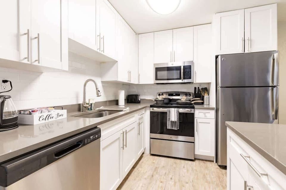 Bright and modern kitchen featuring crisp white cabinetry, stainless steel appliances including a refrigerator, stove, microwave and dishwasher. The sleek gray countertops complement the light wood flooring and provide ample workspace