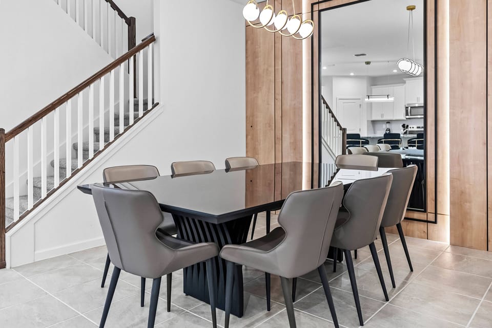 Join us at the sleek black dining table surrounded by stylish gray chairs, perfect for lively conversations. Enjoy the natural light from a large mirror that enhances the space, beautifully complemented by warm wooden accents in.