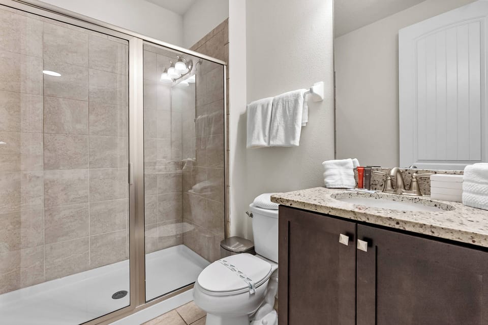 Experience a bright bathroom with sleek granite countertops and dark wood cabinetry for a chic contrast. Enjoy the spacious glass shower and feel pampered with soft towels and neatly arranged toiletries for your comfort.