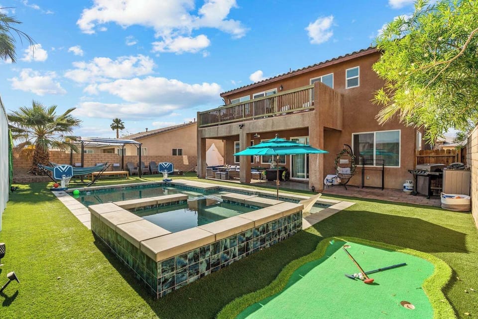 Modern two-story home with a beautiful backyard featuring a rectangular swimming pool with spa, artificial turf, a putting green, and outdoor entertaining areas under a bright blue sky with palm trees.