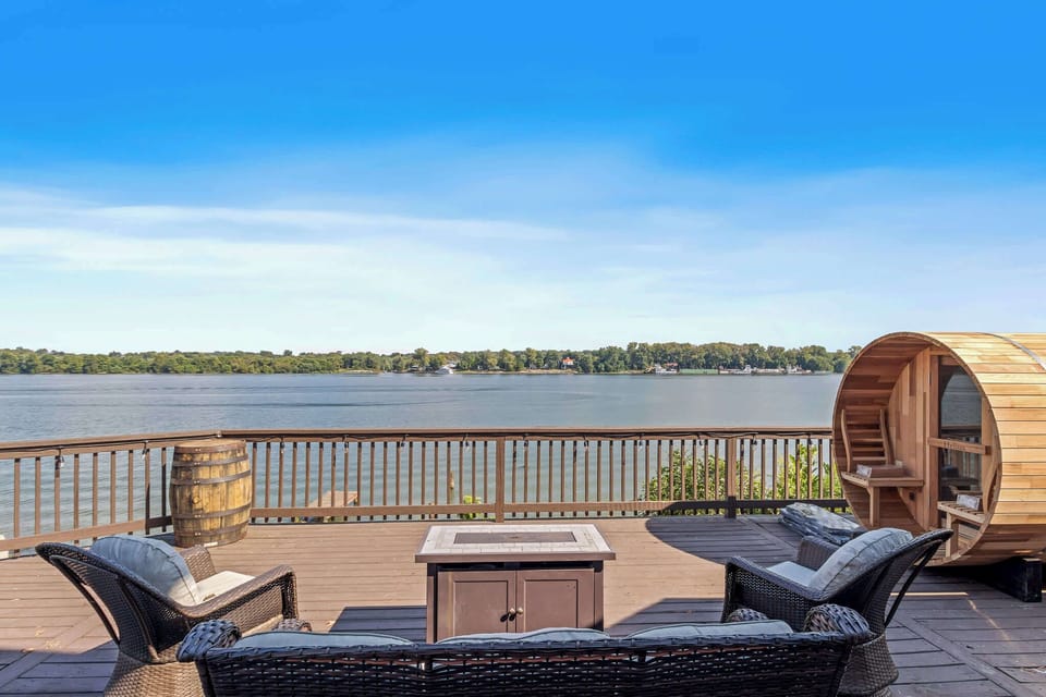 Take in the panoramic waterfront views from an expansive outdoor wooden deck, furnished with a wicker sofa lounge set surrounding a central fire table, a rustic wooden barrel side table, and a wooden outdoor barrel sauna.