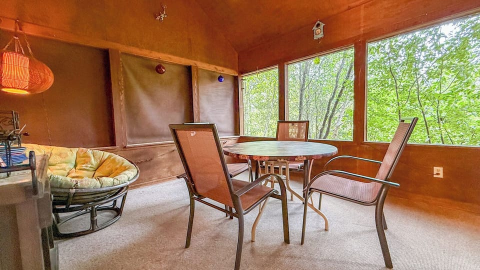 Step into your sunlit retreat where warm wood tones and floor-to-ceiling windows frame lush greenery right outside your door. Gather around the table or sink into the cozy papasan chair—your peaceful escape awaits.