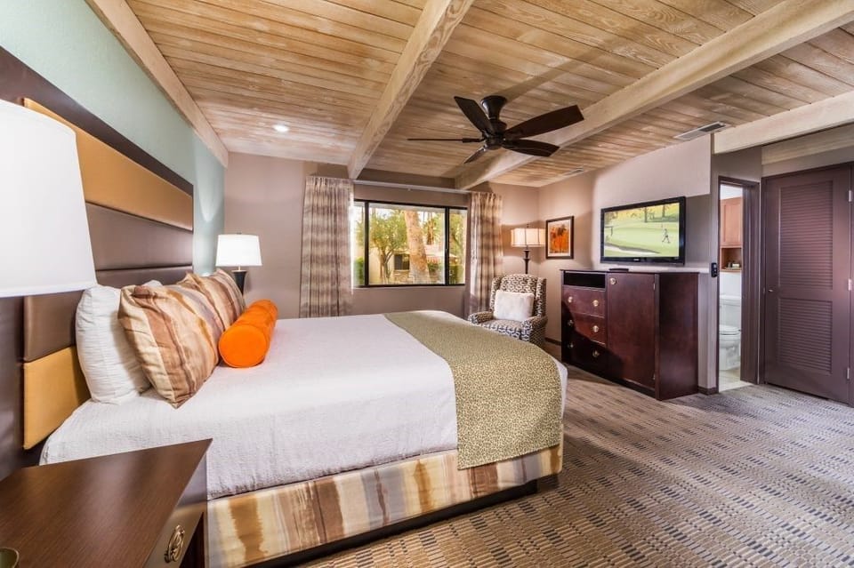 The serene bedroom showcases a plush king-sized bed with colorful pillows, a ceiling fan, and large windows that frame views of the picturesque surroundings.