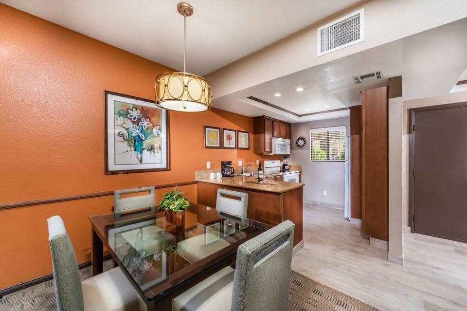 The elegantly designed dining area boasts a stylish glass table, surrounded by comfortable seating and accented by vibrant artwork against a warm orange wall.