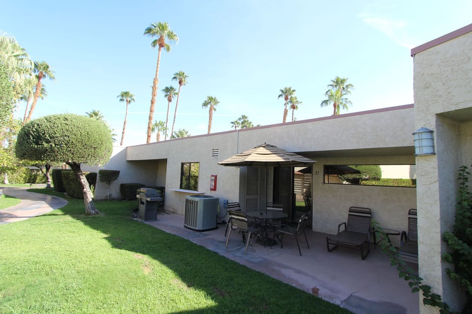 The exterior view reveals a relaxing patio surrounded by well-manicured landscaping, complete with outdoor seating and palm trees in a picturesque setting.
