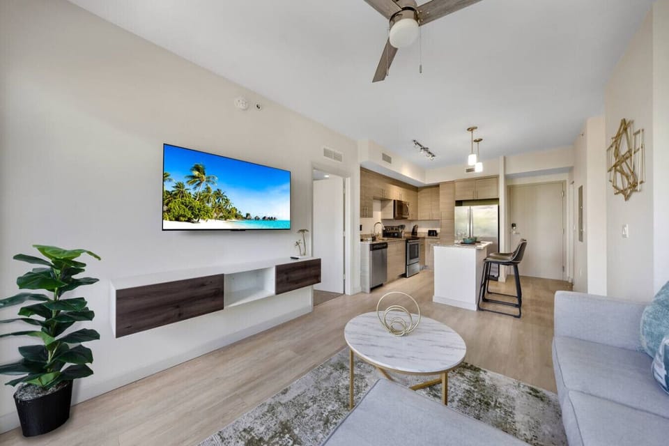 Open Living and Kitchen Space-Contemporary living area with wall-mounted TV, modern décor, and seamless flow into a fully equipped kitchen with island seating. Bright, open design ideal for relaxing or entertaining.