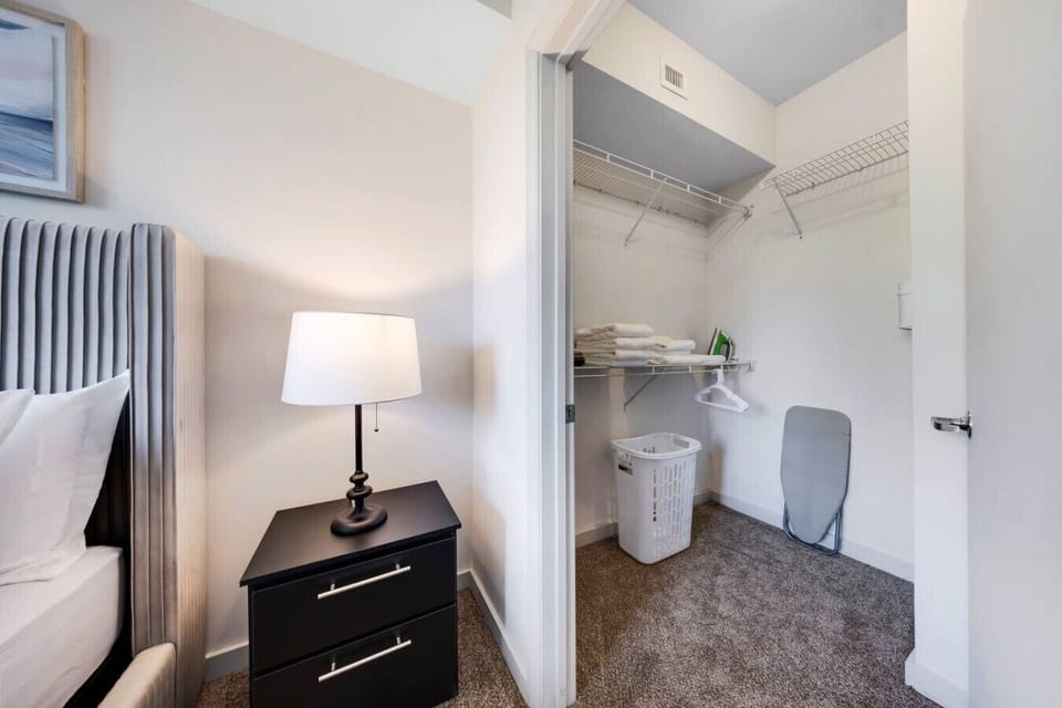 Bedroom with Walk-In Closet-Convenient bedroom setup with bedside table and lamp, leading to a spacious walk-in closet. Equipped with shelves, linens, hangers, laundry basket, and ironing board for your stay.