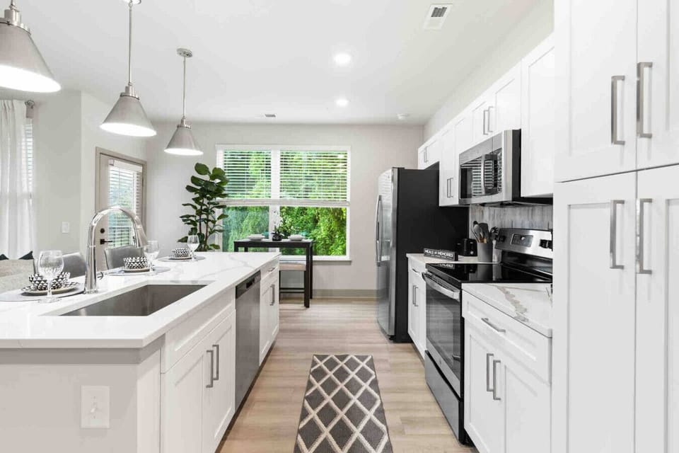 Modern Kitchen with Natural Light- Bright kitchen with sleek white cabinets, stainless steel appliances, and a spacious island with seating. Large windows bring in greenery and light, creating a fresh, inviting space for cooking and dining.